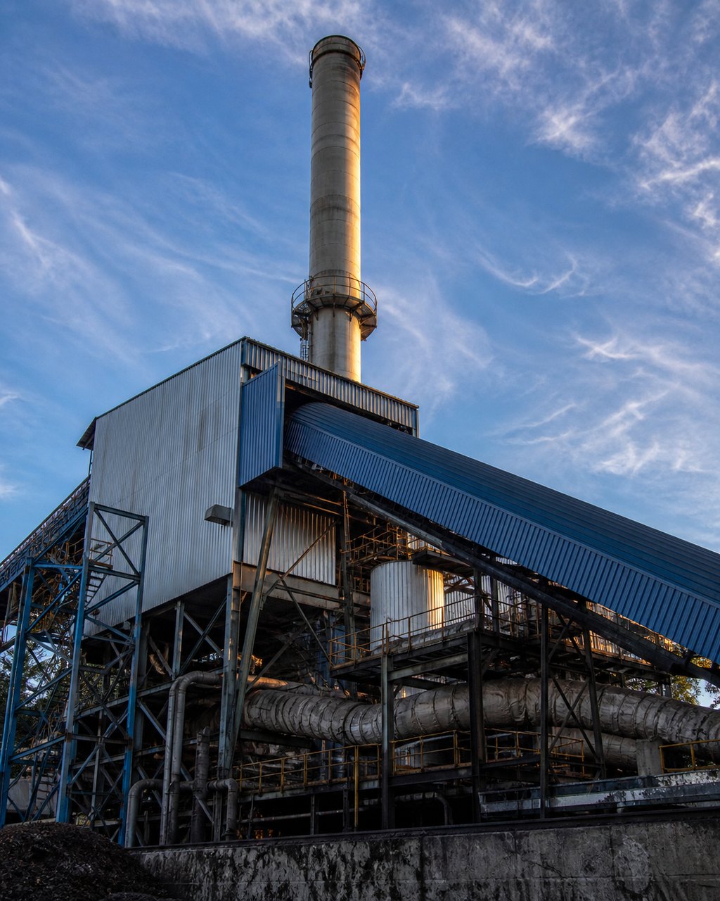 A mill boiler house with its tall stack, blue-roofed sheds and pipework below, seen from outside against a bright sky.