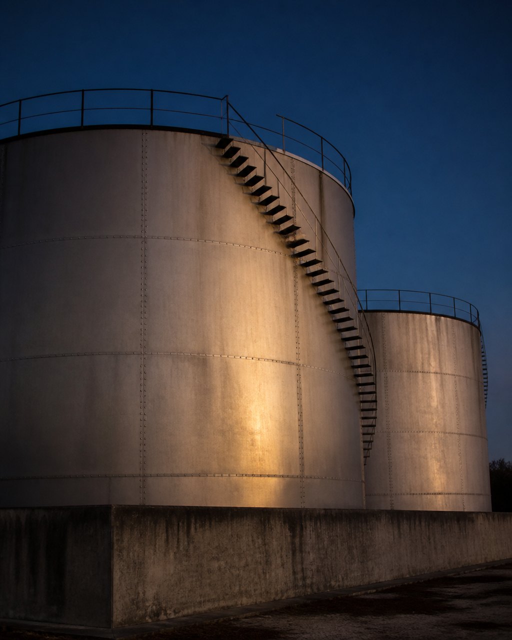 Crude palm oil storage tanks at dusk, a spiral access stair climbing the nearest shell, warm low light on the steel.
