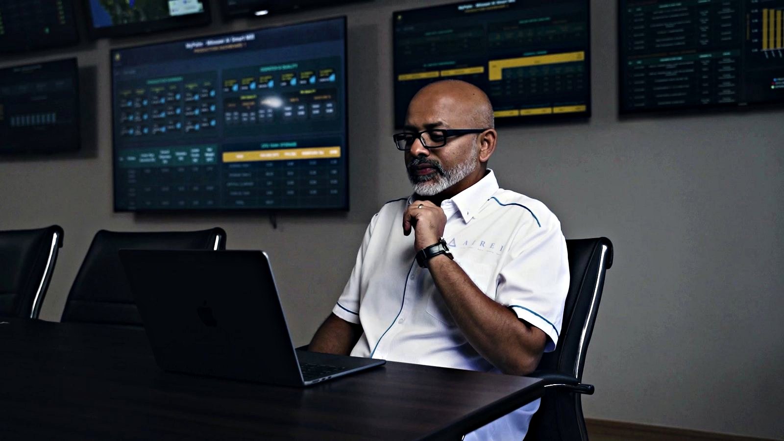 A man in a white AIREI-branded shirt sits at a dark table with an open laptop, chin resting on his hand, reading. Behind him a wall of wide screens carries dark dashboards of tiled panels and tables in amber and pale blue, too small to read here.