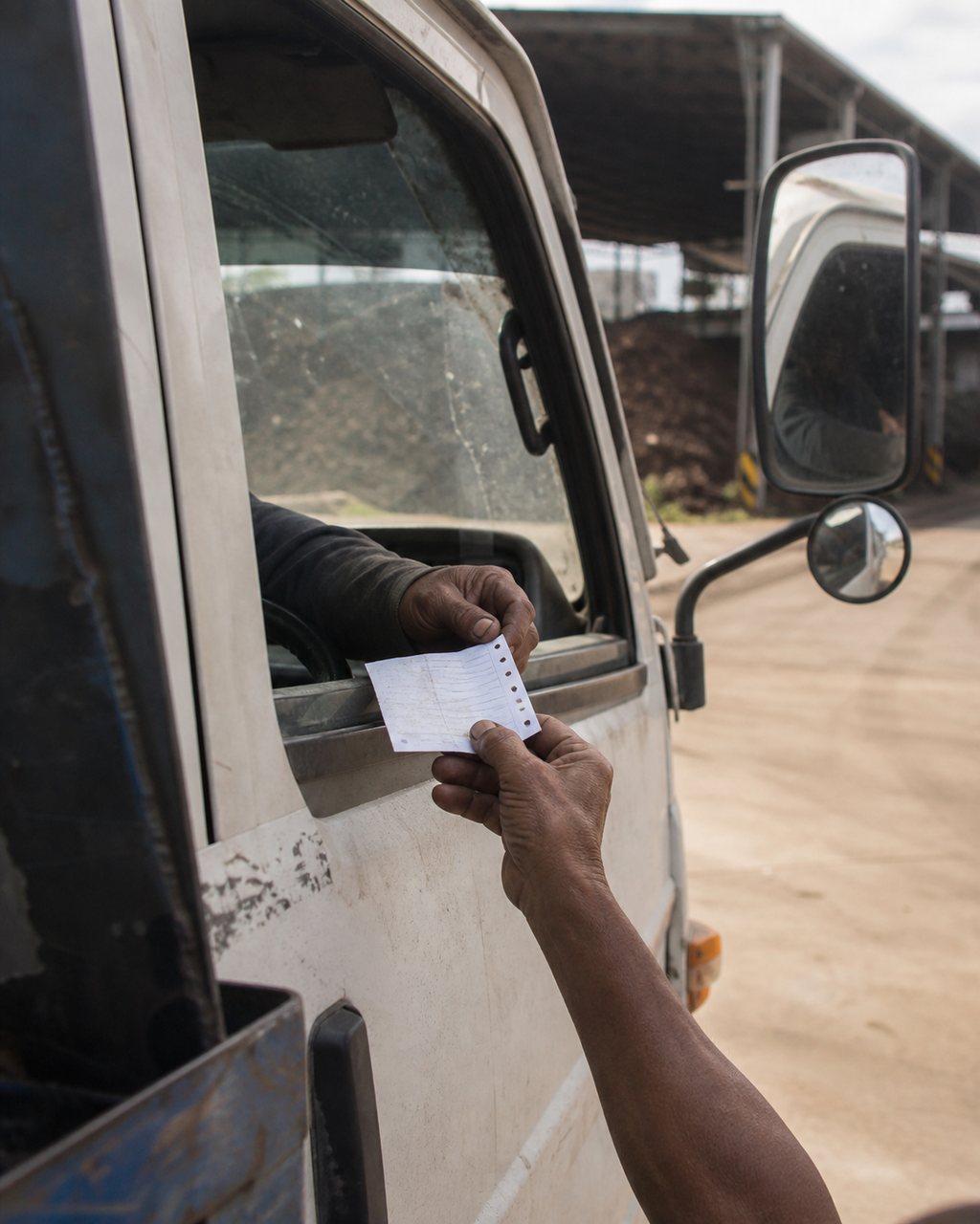 A weathered hand passing a small printed paper docket through the open window of a lorry cab to the driver, whose hand reaches out to take it. Dusty windscreen, mill sheds out of focus behind. No face is visible.