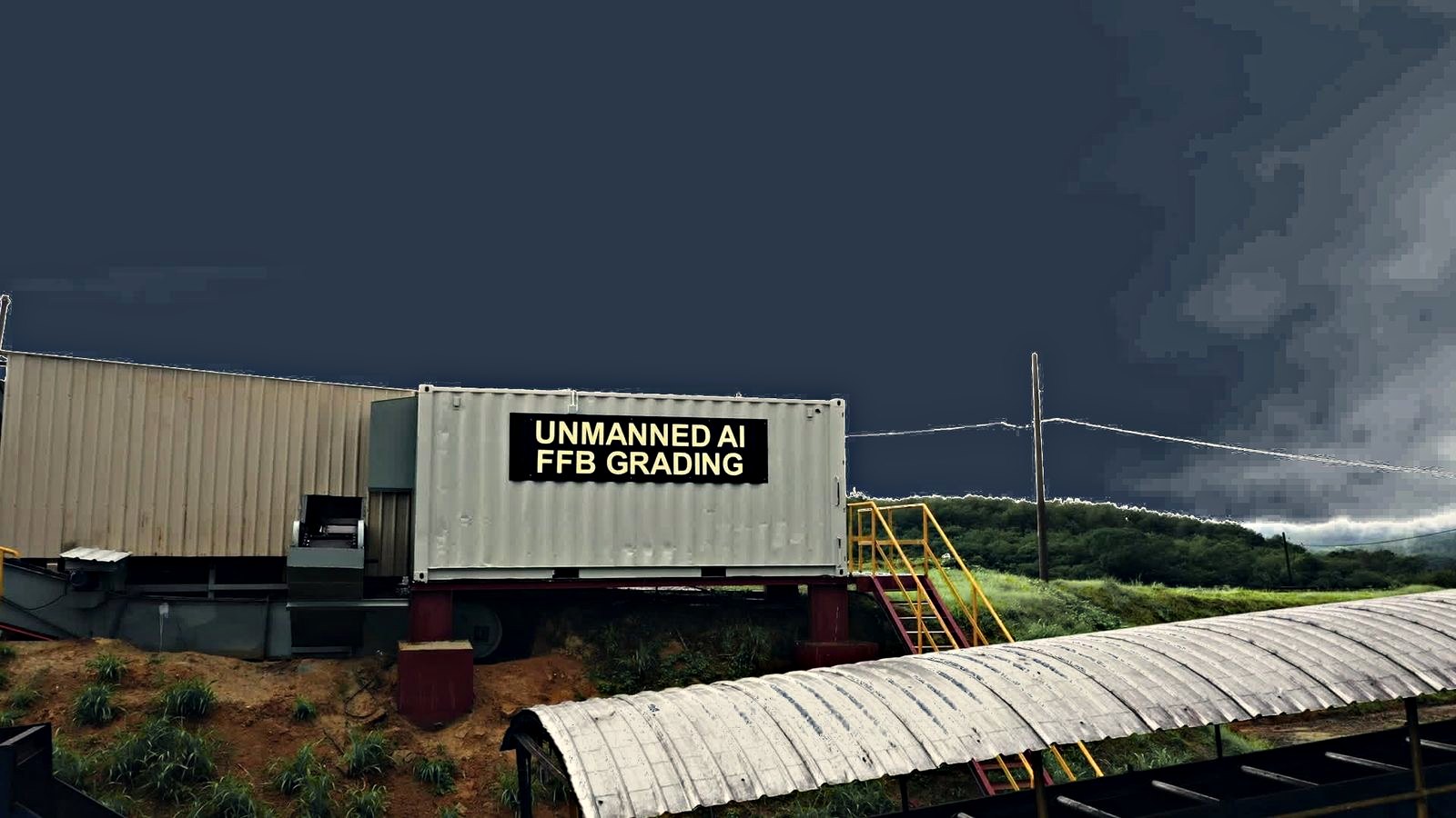 Unit penggredan BTS tanpa pekerja AIREI &mdash; sebuah kontena kapal dinaikkan atas tapak keluli merah, membawa papan tanda hitam bertulis UNMANNED AI FFB GRADING, dengan tangga akses kuning di hujung kanan dan langit ribut di belakangnya.