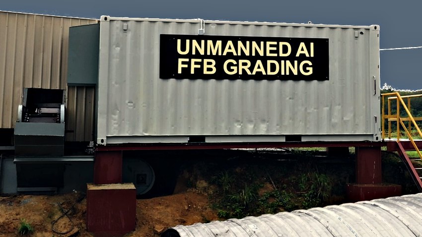 The AIREI unmanned FFB grading unit seen close from the side, filling the frame — a shipping container raised on red-oxide steel legs and a dark roller housing, carrying a black sign lettered UNMANNED AI FFB GRADING in gold capitals across its flank. The intake machinery and the mill shed run off to the left, a yellow access stair stands at the right, and the corrugated roof of a conveyor gallery clips the bottom-right corner.