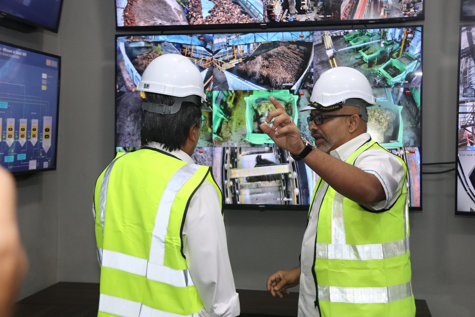 Two men in hi-vis vests and white hard hats standing at the mill data room screens, one gesturing at a live process display.