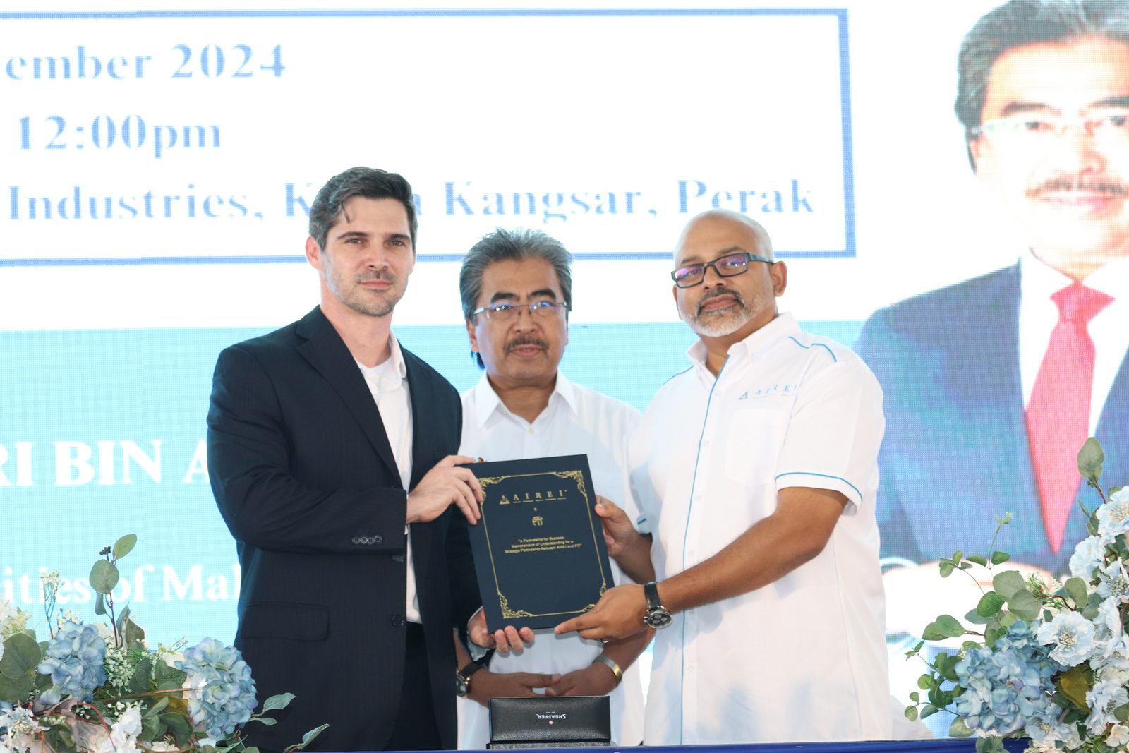 Three men standing for a photograph, the centre one holding a dark presentation folder, against a printed event backdrop.