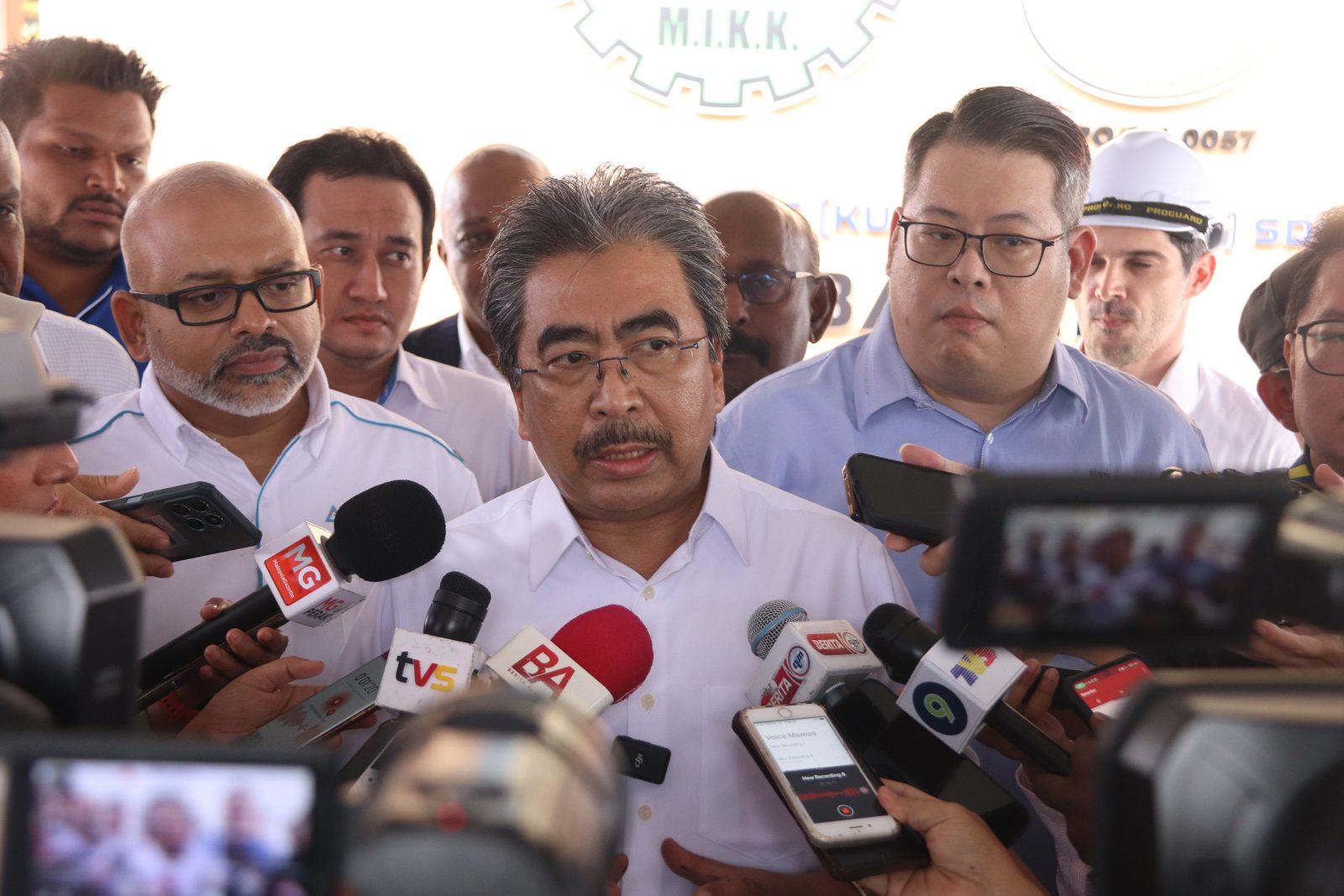 A man speaking into a dense cluster of broadcast microphones at a press conference, reporters crowded around him.