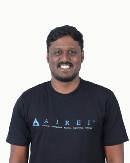 Potret studio di atas latar putih suam yang rata: seorang lelaki berambut kerinting pendek dengan misai dan janggut yang dikemas, tersengih ke arah kamera, memakai baju-T hitam bercetak tanda kata AIREI biru.