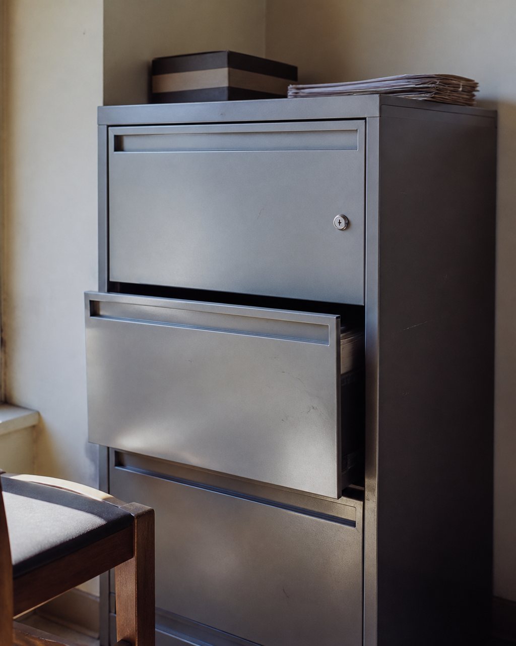 A grey steel document cabinet in a quiet office, one drawer very slightly open, a small keyhole lock visible in soft daylight.