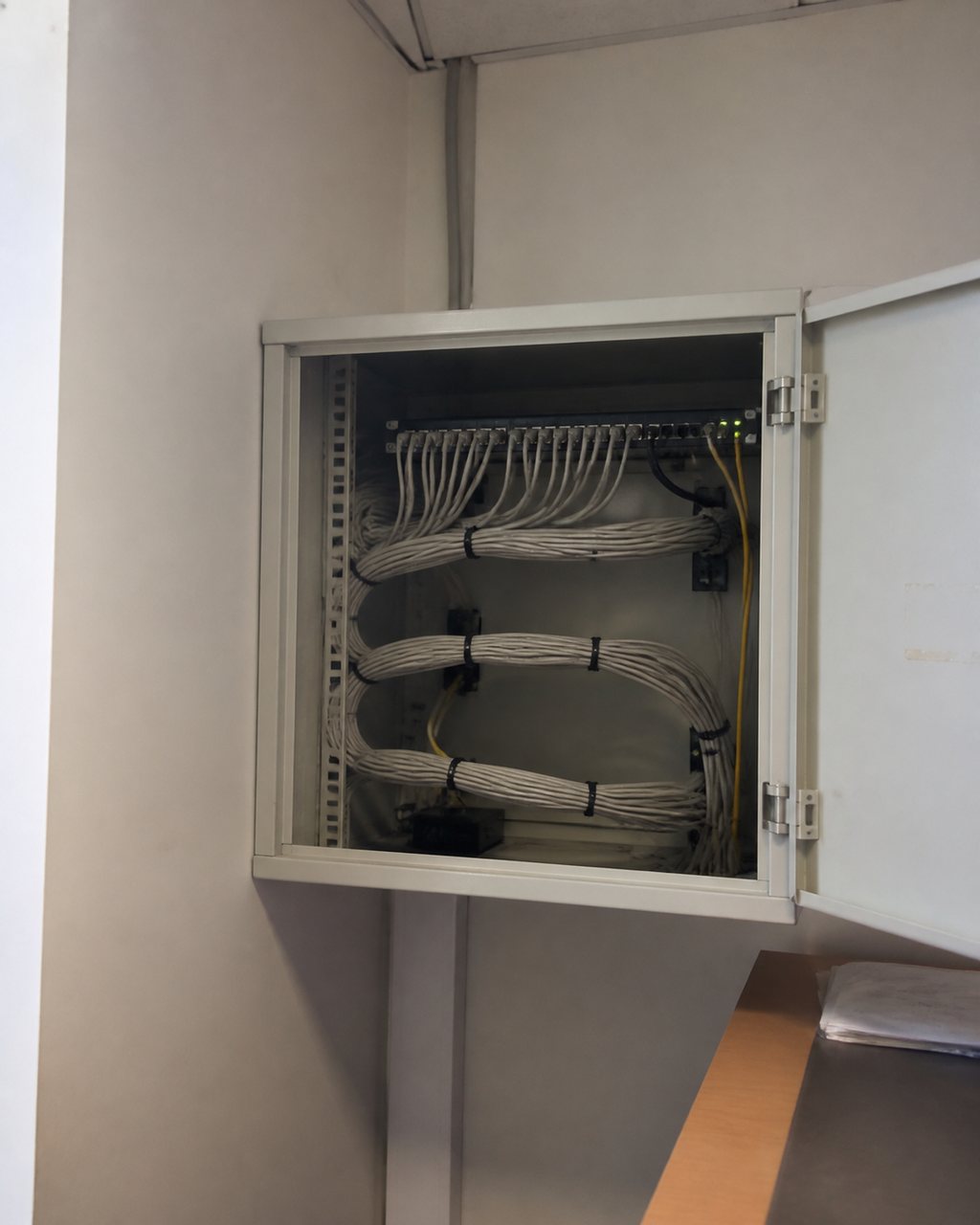 A small comms cabinet in the corner of an estate office with its door open: a patch panel, neat looms of network cable and two green indicator lights.