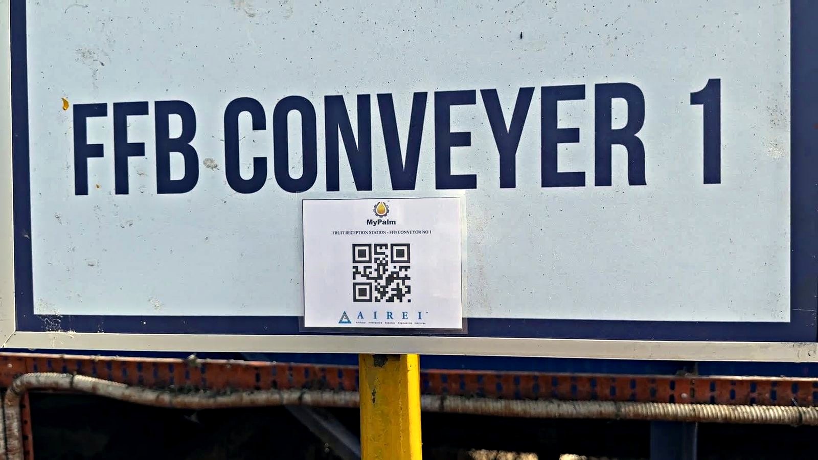 A weathered white steel sign with a dark navy border, hand-painted in tall navy capitals reading FFB CONVEYER 1. Taped to the lower centre of the sign is a small printed white label headed with the MyPalm droplet mark, the line FRUIT RECEPTION STATION - FFB CONVEYOR NO 1, a square QR code, and the AIREI logo beneath it. Below the sign, a yellow painted post and the grey mesh guarding of the conveyor run across the bottom of the frame.
