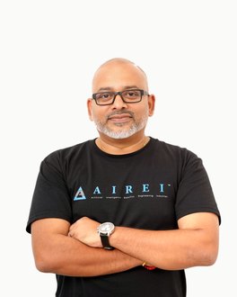 Potret studio di atas latar putih suam yang rata: seorang lelaki berkepala botak dengan janggut putih pendek dan cermin mata berbingkai hitam segi empat, berpeluk tubuh, memakai baju-T hitam bercetak tanda kata AIREI dan sebuah jam di pergelangan tangan kirinya.